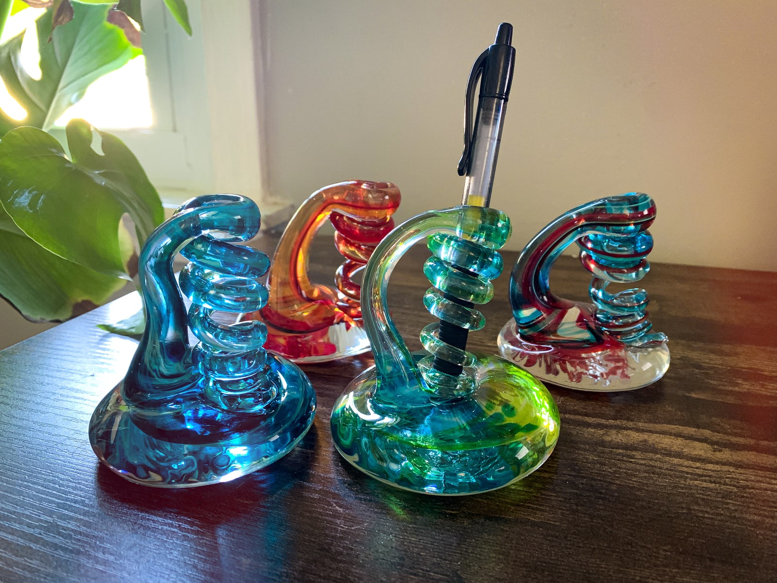Four blue, green, and red curled glass pen holders beside a leafy plant