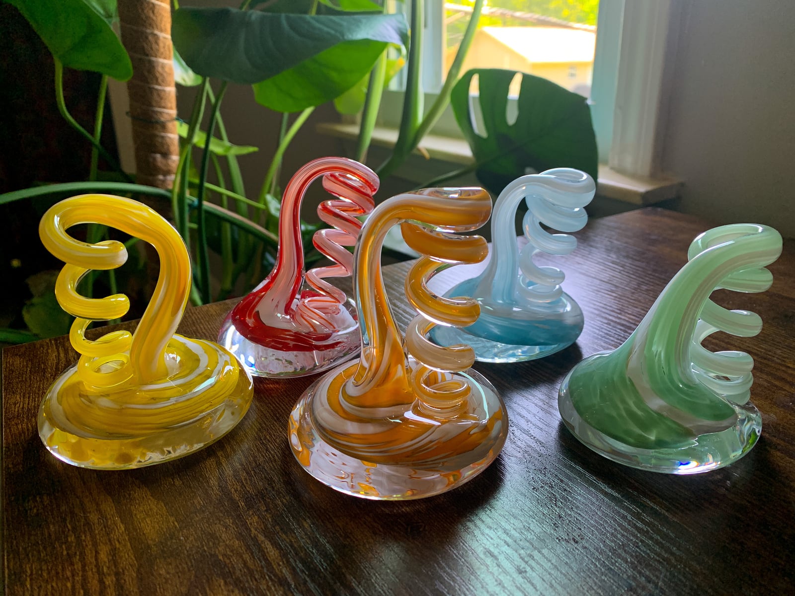 Five colourful glass sculptures with looping curled shapes beside a leafy plant
